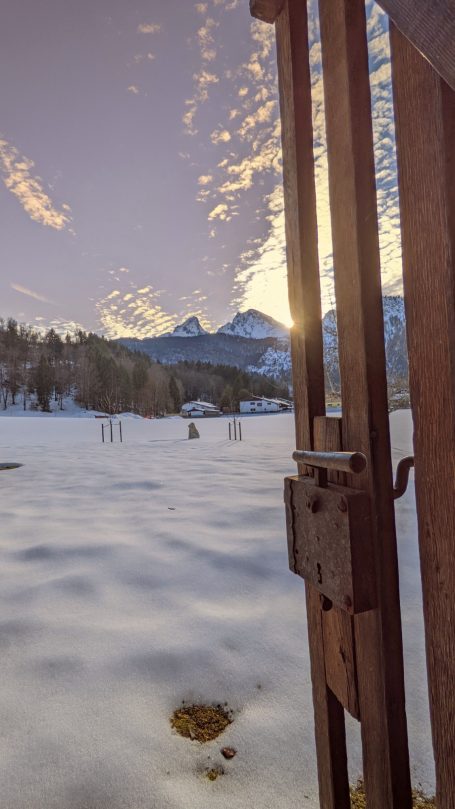 Blick durch ein Tor auf schneebedeckte Landschaft und Berge bei Sonnenuntergang.