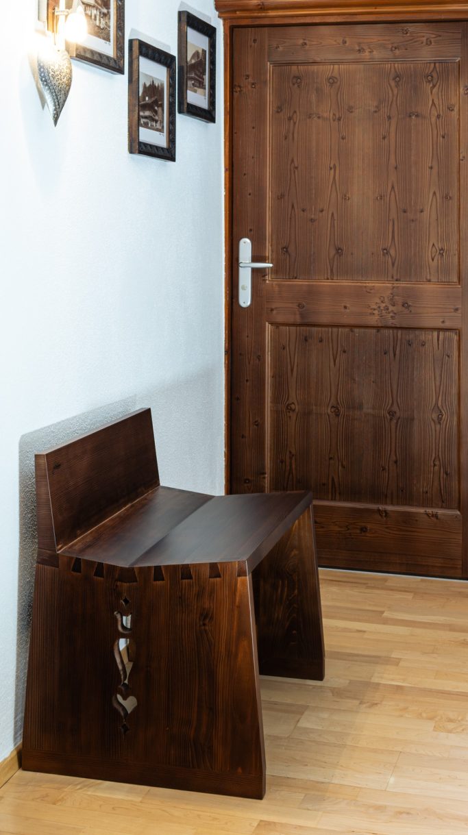 Holzbänkchen in einem Flur neben einer Holztür und gerahmten Bildern an der Wand.