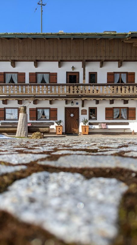 Bauernhaus mit Holzverkleidung und blauen Fenstern, umgeben von Steinpflaster.