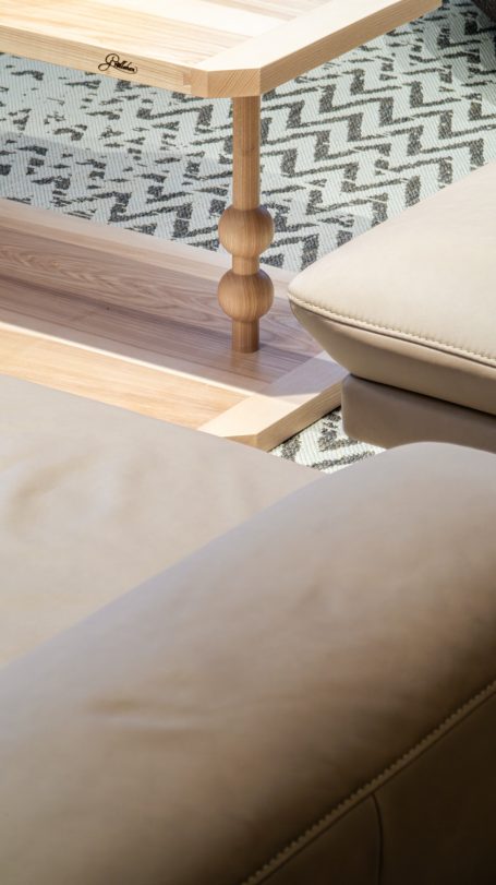 Holztisch mit einem runden Bein, Sicht auf eine beige Couch und ein gemustertes Teppichmuster.