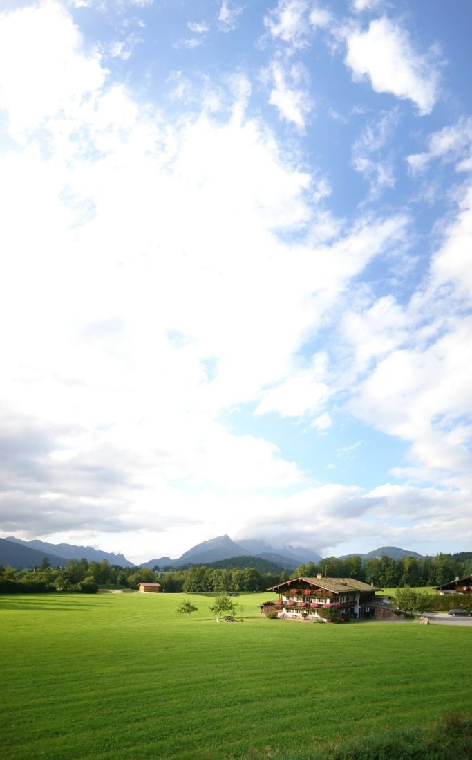Landschaft mit einem kleinen Haus, umgeben von Wiesen und Bergen unter einem bewölkten Himmel.