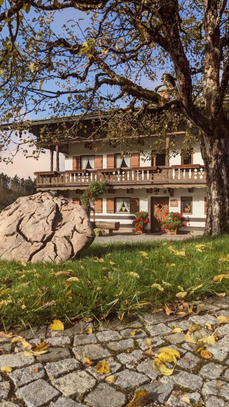 Traditionelles Haus mit Balkon, umgeben von Bäumen und buntem Laub. Großer Stein im Vordergrund.