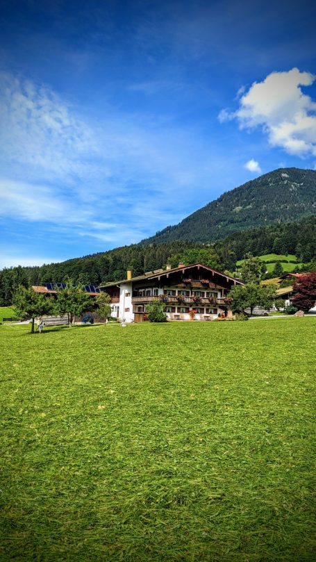 Landschaft mit grünem Feld, traditionellen Gebäuden und Bergen im Hintergrund.