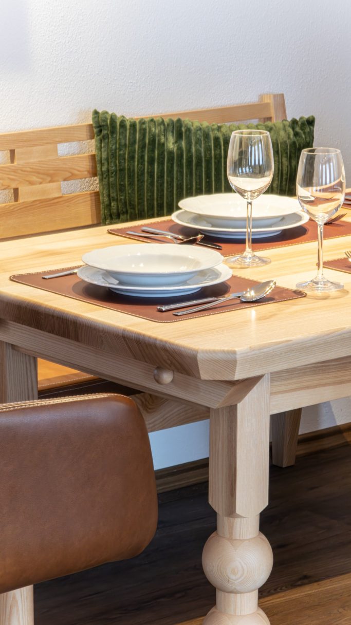 Holztisch mit sauberem Tischgedeck, Gläsern und einem Kissen im Hintergrund.