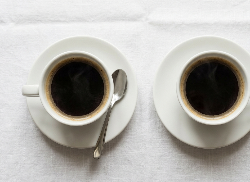 Zwei Tassen Kaffee auf einem Tisch, jeweils mit einem Löffel daneben.