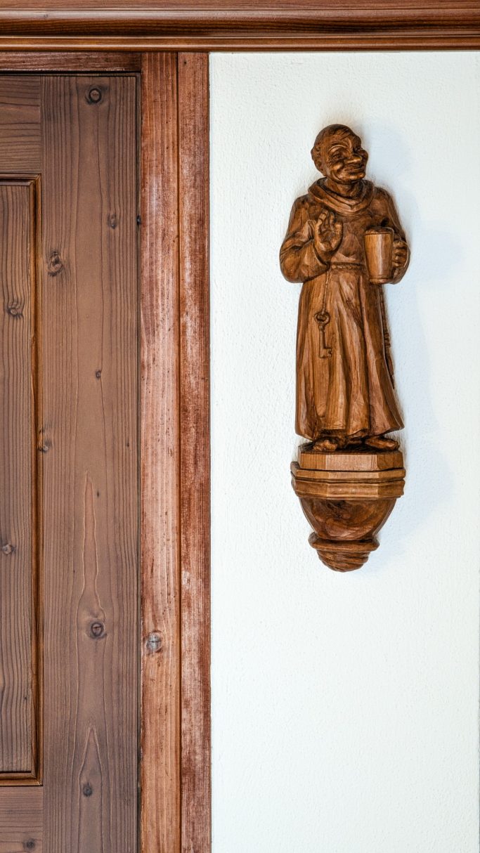 Holzfigur eines heiligen Mannes, an einer weißen Wand neben einer Holztür angebracht.